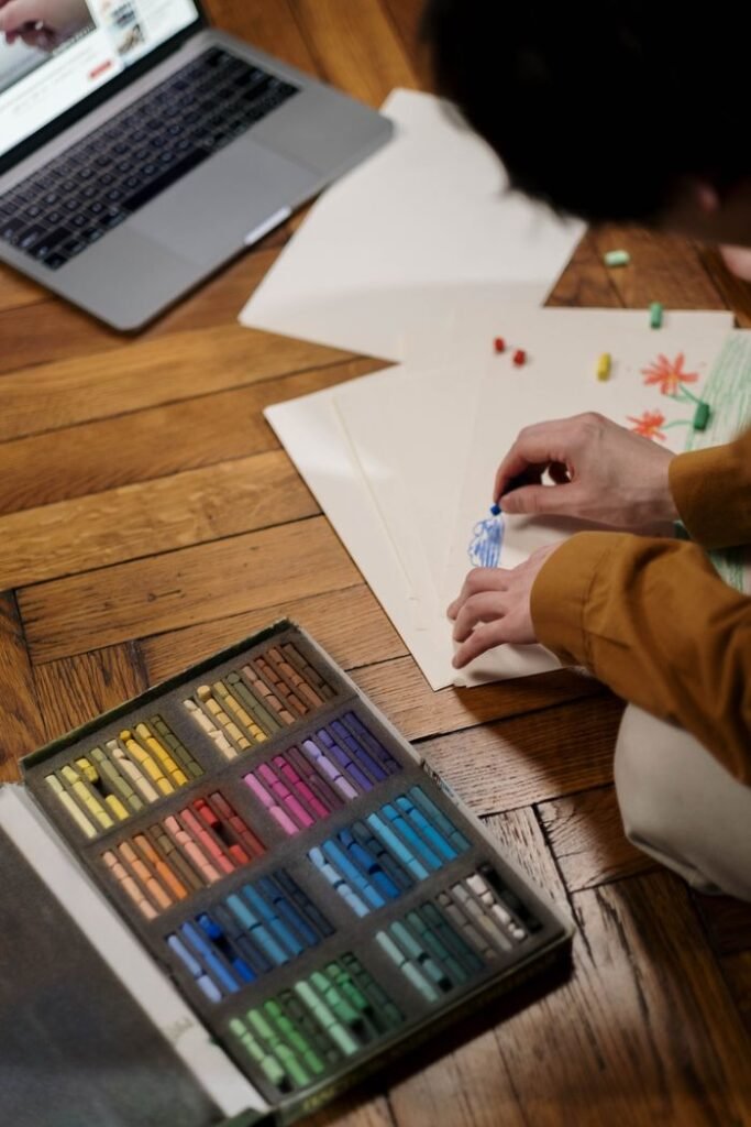 Adult engaged in online art class, using oil pastels on wooden floor.